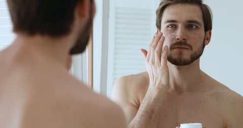 Attractive shirtless metrosexual young guy applying moisturiser cream on face looking in mirror. Handsome bearded man holding creme jar doing morning skin care beauty routine. Male skincare concept