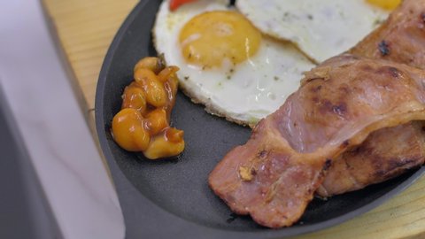 Full English Breakfast served in a pan. Fried eggs, beked beans, tomatoes, champignons, crispy bacon, sausages and toast. Placed on stone background. Top view with copy space.