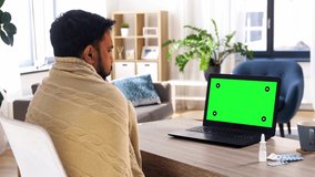 healthcare, technology and people concept - sick indian man in blanket with thermometer having video call on laptop computer with chroma key green screen at home