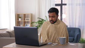 healthcare, technology and people concept - sick indian man in blanket with thermometer having video call on laptop computer at home