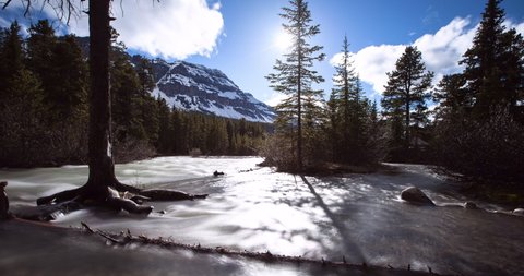 Time lapse shot of creek flowing by forest and mountains against sky on sunny day - Banff, Canada