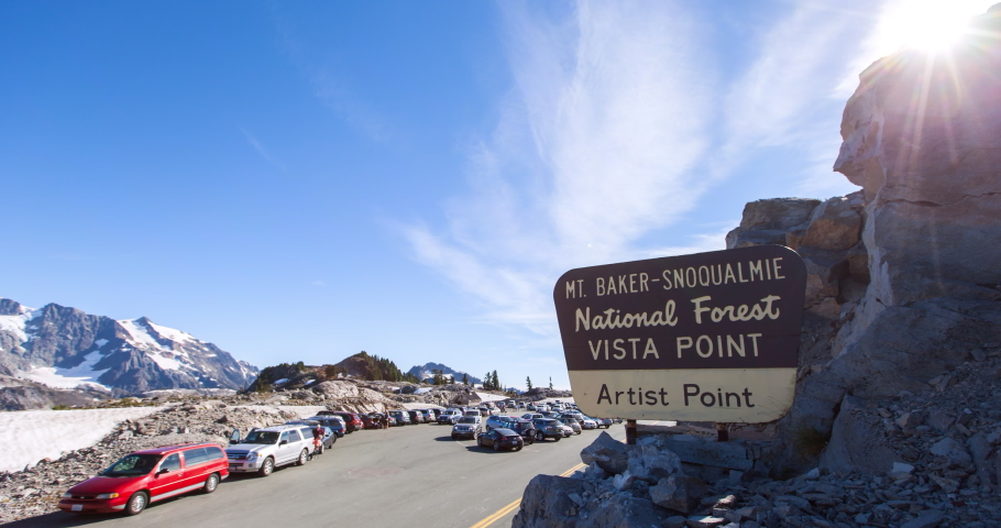 Lockdown time lapse shot of signboard on rocks by vehicles and tourists against sky during sunny day - Mount Baker, Washington