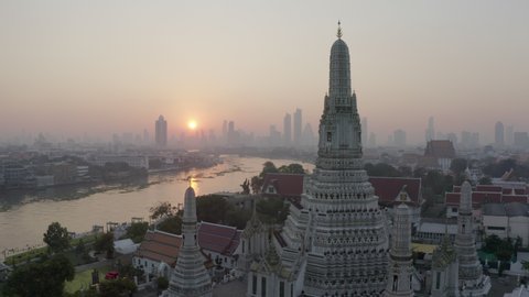 Aerial shot of Buddhist temple by river in city at sunset, drone flying from famous landmark against sky - Bangkok, Thailand