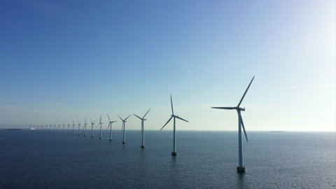 Aerial panning shot of wind turbines in sea against blue sky, drone flying over water on sunny day - Copenhagen, Denmark
