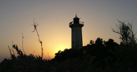 Low angle panning shot of lighthouse amidst silhouette plants and trees against sky during sunset - Camargue, France