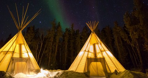 Time lapse shot of illuminated teepees on snow by trees against sky at night - Northwest Territories, Canada