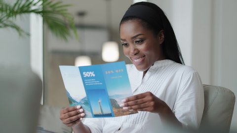 Happy African American woman sitting on sofa at home and planning holiday trip. Black people reading travel flyer advertising promotional discount for vacations