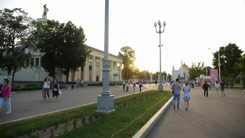 Moscow/Russia - JUNE 01.2019: Young couple walks on main avenue of Vdnkh park in Moscow. Dolly shot of  people walk along the alley where a Magnificent lawn and green shrubs in sunset. 