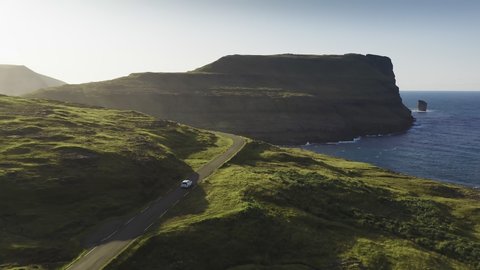 Car driving along coastal road view on wild green island at sunset.Cinematic rear view approaching.Faroe Island road along beautiful coast. Faroe island, Denmark. Forward aerial flight
