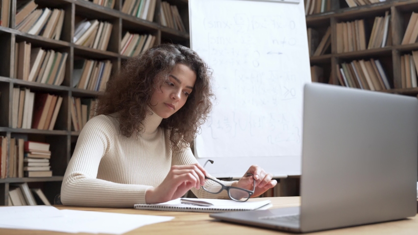 Young hispanic school math teacher sitting at workplace, portrait. Confident smiling latin female professional tutor looking at camera at library desk with laptop. Teaching, online education concept. Royalty-Free Stock Footage #1052462404
