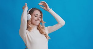 Happy woman enjoying music in wireless headphones and dancing, blue studio background