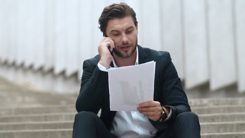 Upset businessman talking on smartphone in modern city. Portrait of aggressive male manager analyzing diagrams outside. Closeup angry business man throwing documents on floor at street