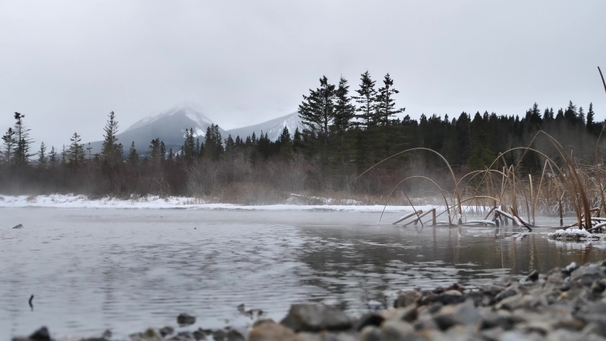 Low angle shot with steam above Banff Lake. Mountains and trees are in the background with rocks and pebbles in the foreground.