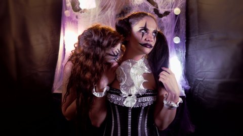 Girl with Halloween makeup is sitting in front of a mirror. Makeup of the actor. The girl in the image of the devil's Bride comes up and bites the neck of the clown girl.