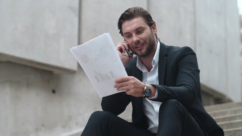 Low view smiling business man calling on mobile phone in modern city. Closeup positive male executive looking at business documents outdoors. Businessman waving head yes. Man sitting on stairs