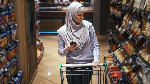 Portrait of young beautiful muslim woman in hijab using phone and choosing food in the supermarket, slow motion