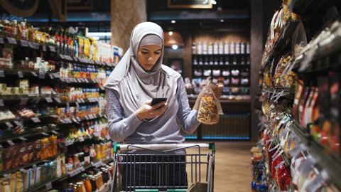 Portrait of young beautiful muslim woman in hijab using phone and choosing food in the supermarket, slow motion