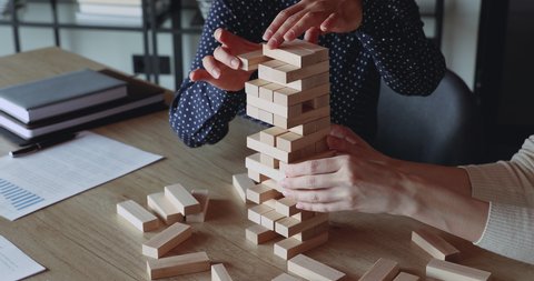Two business women play build game in office. Female business partners hands take blocks out of wooden tower. Risky solutions in company building strategy plan, help in teamwork concept. Close up view