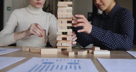 Female partners taking wooden bricks out of tower playing build game on office table. Business insurance, effective risk strategy, building economic stability, credit investment concept. Close up view