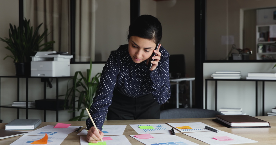 Young busy indian business woman working on project paperwork talking on the phone. Ethnic female manager making business call on mobile writing tasks on sticky notes working alone in office. Royalty-Free Stock Footage #1052490031
