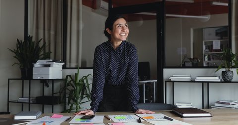Happy young indian business woman standing at workplace table with papers and sticky notes looking at camera. Smiling ethnic female manager posing in office. Corporate planning concept. Portrait.