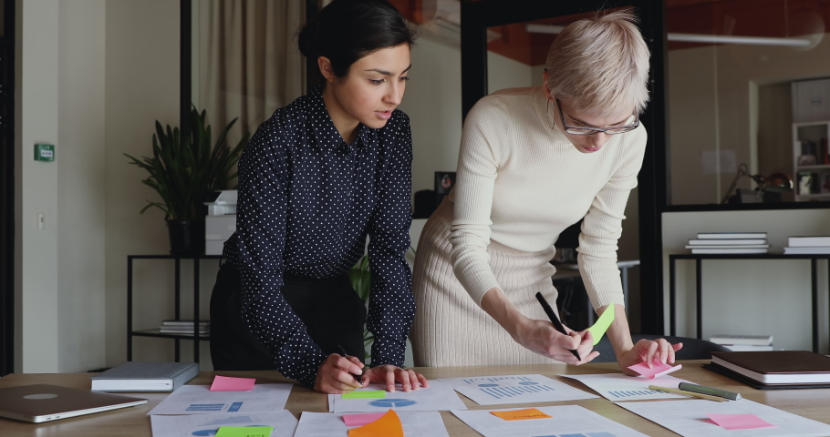 Diverse young business women brainstorm on project plan paperwork applying sticky post it notes on table. Two indian and caucasian coworkers discuss business strategy put stickers at office meeting. Royalty-Free Stock Footage #1052490046