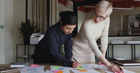 Diverse young business women brainstorm on project plan paperwork applying sticky post it notes on table. Two indian and caucasian coworkers discuss business strategy put stickers at office meeting.