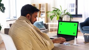 healthcare, technology and people concept - sick indian man in blanket with medicine pills having video call on laptop computer with chroma key green screen at home