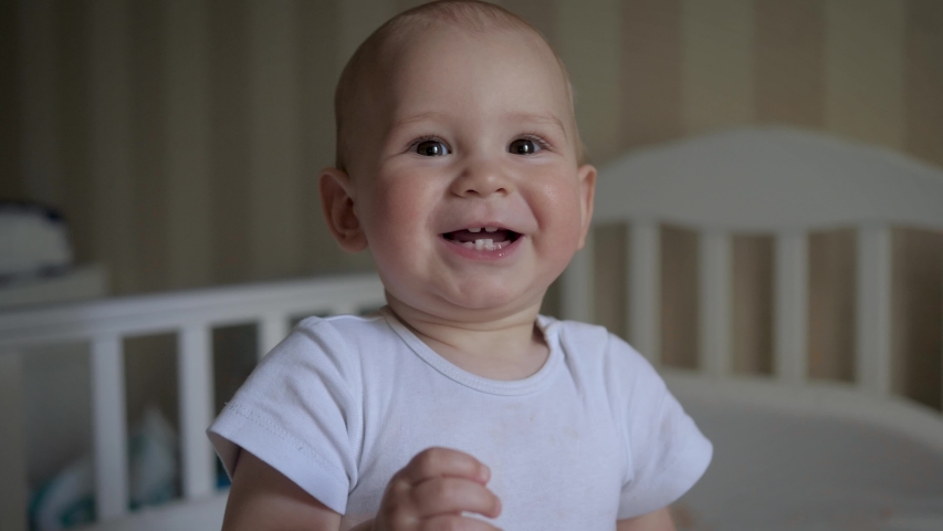 Close up portrait of little baby who learned to stand. Looks at camera and smiles cheerfully, at the child is growing teeth, against the background of a baby cot in the room. Slow motion. Royalty-Free Stock Footage #1052492863