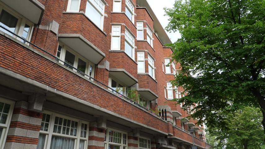 Low angle shot of brick building, multi-unit apartment house at Amsterdam. Five floor apart house with balcony and many bay windows, green tree crown against Royalty-Free Stock Footage #1052493736