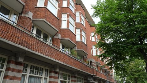 Low angle shot of brick building, multi-unit apartment house at Amsterdam. Five floor apart house with balcony and many bay windows, green tree crown against