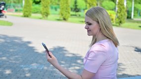 Video Caucasian blonde girl talking on the phone in a park outdoors sitting on a bench in spring sunny weather.