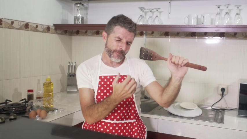 SLOW MOTION singing in kitchen - young cool and attractive man in red apron singing song and dancing crazy happy holding spoon as microphone having fun cooking carefree