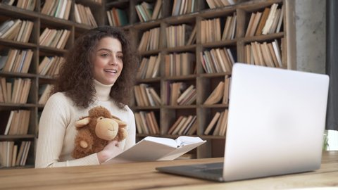 Smiling hispanic woman teacher reading book playing with toy during virtual online kindergarten or school children class looking at laptop webcam teaching kids in distance online application concept.