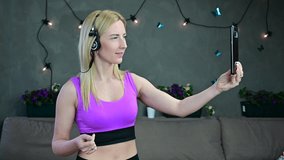 Video caucasian fitness girl blonde cute smiling holds online conversation via smartphone with headphones at home.