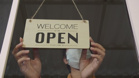 Asian woman wear mask turning Welcome we are open sign on fronton glass door store. Reopening activity after COVID-19, ended the lockdown and quarantine. Restarting again and keep forward concept