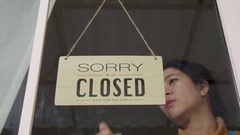 Business owner asian woman turning Sorry closed sign on fronton glass door store. Close activity from COVID-19, the lockdown and quarantine. Starting again and keep save and stay home concept