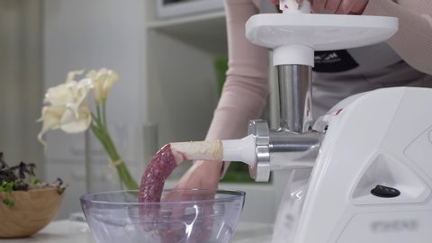 Housewife cooks sausages in traditional way using sausage filler of electric mincer machine. Production of meat delicacies at home. Preparation of raw beef sausages with electric grinder.
