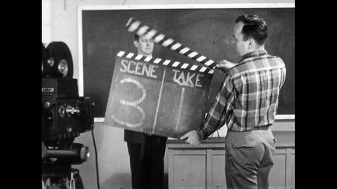 1950s: Director addresses production crew on soundstage. Man in booth waves hand. Camera operators speak. Man operates large clapboard and speaks. Man carries clapper off soundstage.