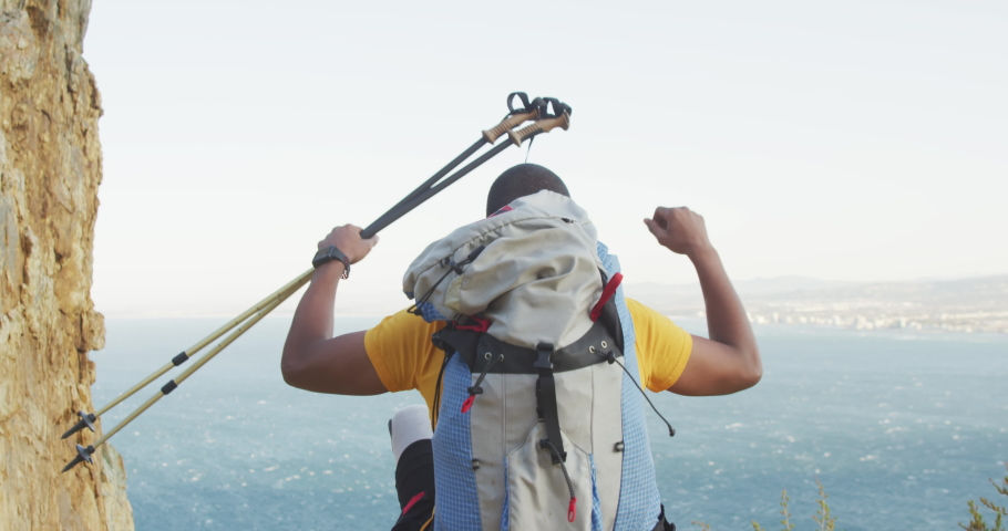 Fit, disabled mixed race man with prosthetic leg, enjoying his time on a trip to the mountains, hiking, standing, raising his hands, holding Nordic walking sticks, on a cloudy day, in slow motion