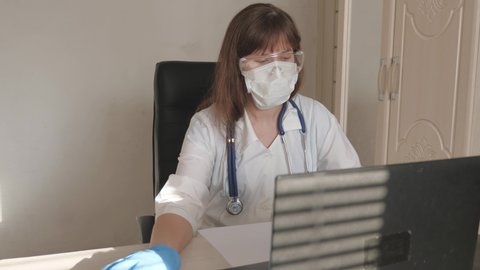 Female doctor in white coat and safety glasses working on laptop, online consultation and conference. Doctor communicates with client using virtual chat. Telemedicine, remote medical care concept.