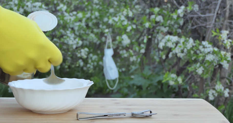 Self-isolation. A man puts canned beans on a plate with a spoon against a background of cherry blossoms and a medical mask hanging on a branch.