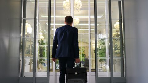 Rear view of young businessman wearing dark suit entering hotel going through glass doors. Man carrying case luggage and briefcase. Tourist concept. 4k footage
