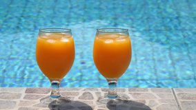 Two bright beautiful orange yellow cocktails juice with ice stand close-up on side pool, against background of pool, blue water. Summer pool party, vacation. Slow motion video.