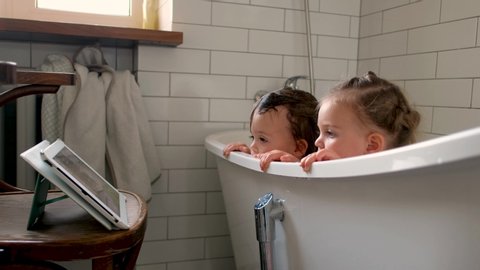 Curious obedient children focusing on screen of digital tablet on stand and watching cartoon while washing in bath in light bathroom with simple design of interior in retro style