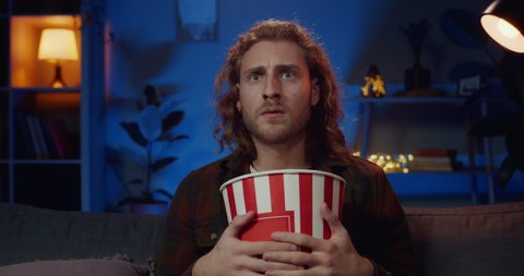 Handsome hipster man making shocked facial expression while watching interesting film. Millennial bearded guy hugging paper popcorn bowl while sitting on couch in front of tv at home