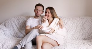 Young couple watching TV on the grey sofa at home in bright living room. Exciting couple interested on tv show eating popcorn