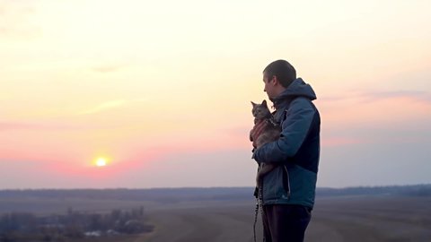 a man with a cat at sunset, a walk in the spring with a pet. the cat is sitting on the man's hand. sunset sky on the horizon.