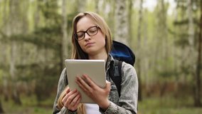 Young caucasian woman with glasses and backpack smiling while holding tablet and walking in the nature