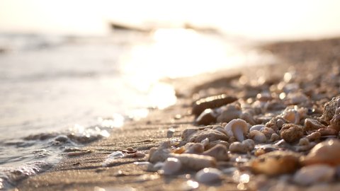 Blured bokeh nature sea beach tropical background in morning sunlight. Shimmering sea wave foams wash up on sand beach with sea shells debris and pebbles sparkling shiny reflected to sun light.